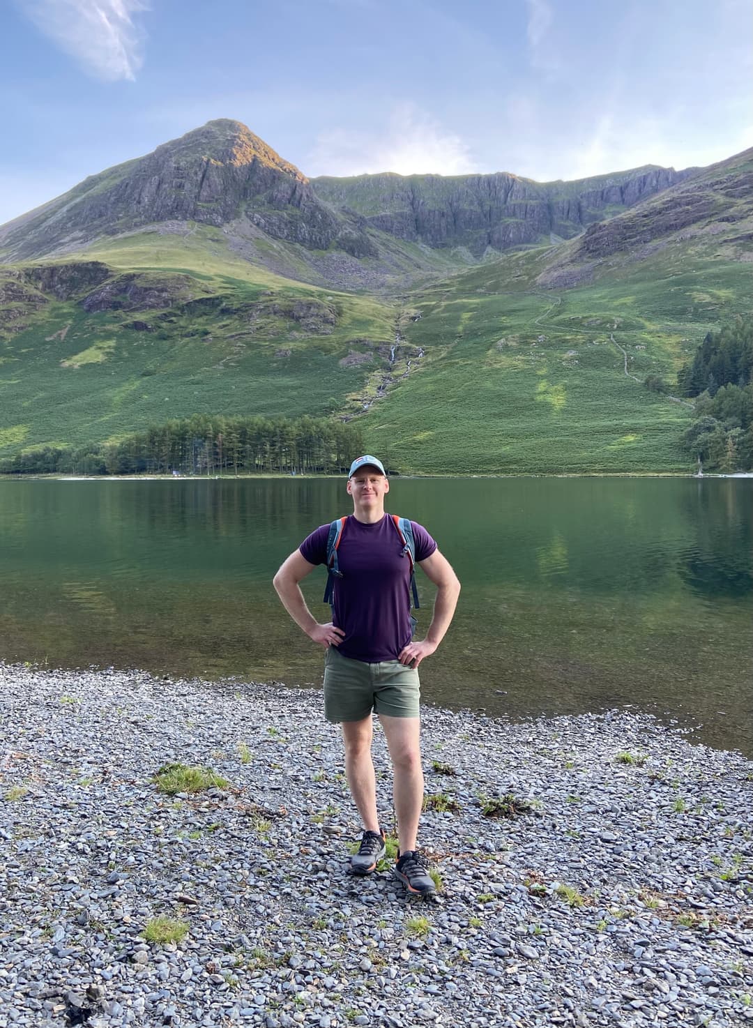 Justinas with a hiking backpack on the stony shore of a lake, mountains behind him